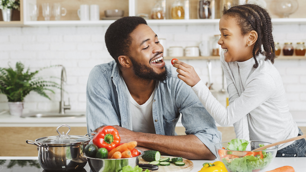 African girl feeding dad in kitchen, giving him cherry tomato for her holistic whole food diet